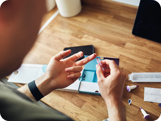 Persona che preleva un campione di sangue dal dito durante un controllo sanitario a casa, con strumenti medici sul tavolo in legno.