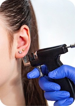 Orecchio di una persona con piercing, mentre una mano con guanti blu e pistola per forare i lobi sta per aggiungere un altro buco