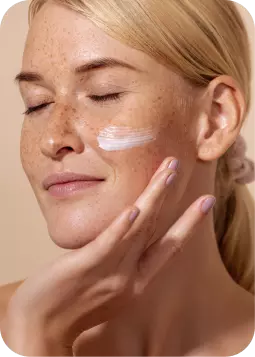 Donna con pelle chiara e lentiggini, applica crema bianca sulla guancia con le dita, in primo piano.