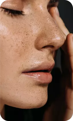 Primo piano di una pelle matura con pori visibili, lentiggini e labbra leggermente lucide, luce neutra su tonalità calde.