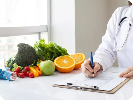 Medico firma una cartella clinica accanto a frutta fresca: arance tagliate, mele, verdure e una confezione di integratori sul tavolo.