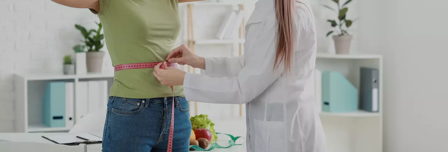 Donna in camice medico misura girovita a una paziente in maglietta verde, con frutta fresca sul tavolo in background.