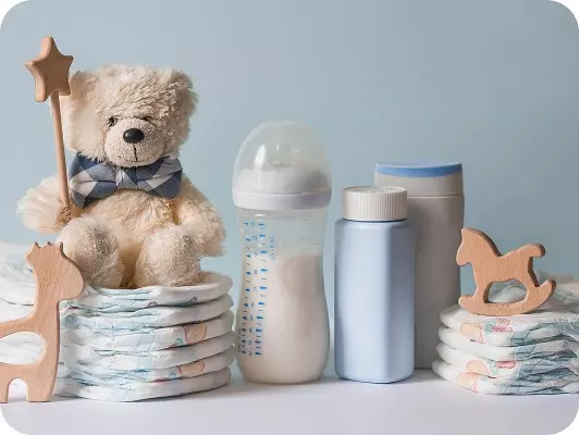 Setup per neonato: peluche orso con bacchetta stellata, biberon, latte in lattine e contenitori azzurri, pannolini impilati e giocattoli in legno su sfondo azzurro.