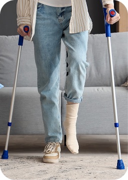 Primo piano sulle gambe di una persona sorretta da due stampelle, una gamba con calzamaglia e gesso, ambiente domestico, neutro.